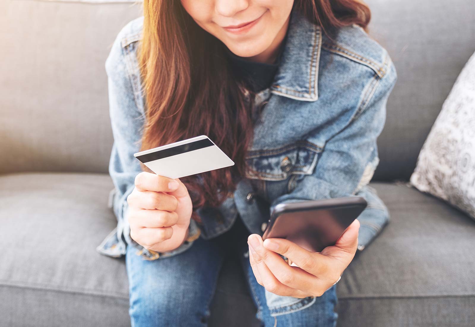 Eine junge Frau sitzt auf dem Sofa, in der einen Hand ein Smartphone, in der anderen Hand eine Bankkarte